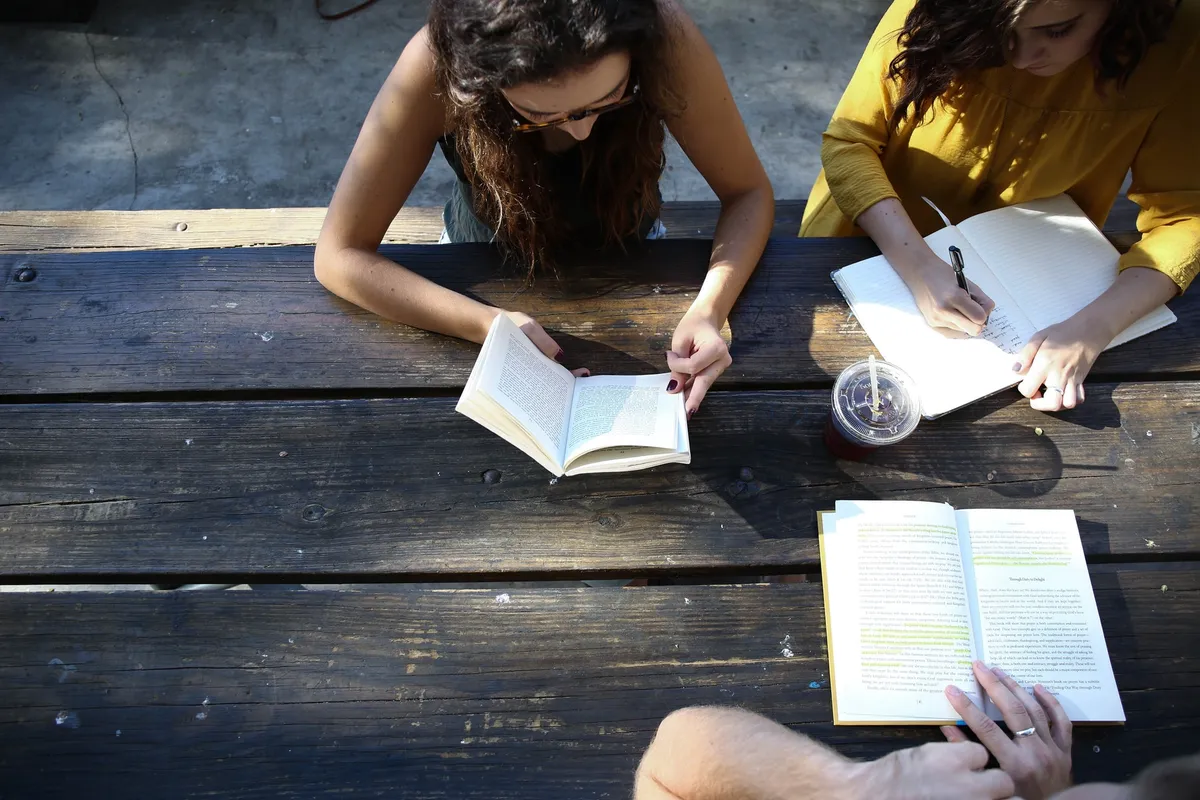 Three students at an outdoor wooden table, one textbook marked up in yellow highlighter.
